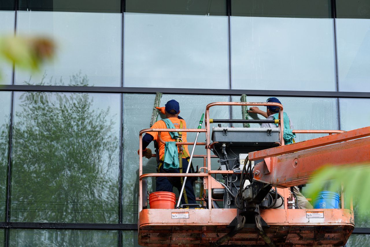 services-02 Deux intervenants nettoient les vitres d’une façade d’immeuble en verre à l’aide d’une nacelle, afin d’améliorer l’esthétique du bâtiment.
